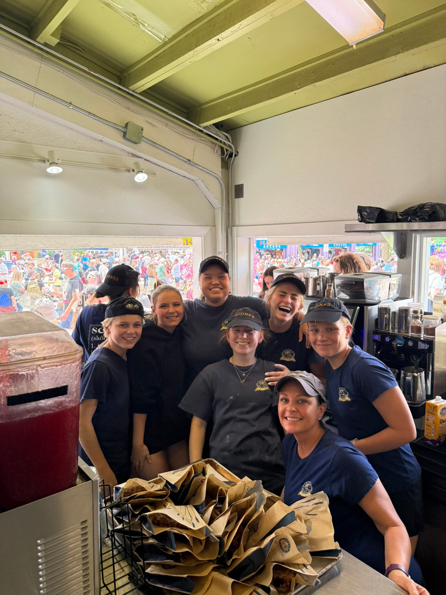 The French Meadow kitchen team smiling together during a busy Fair day, with the fairground crowds visible through the windows